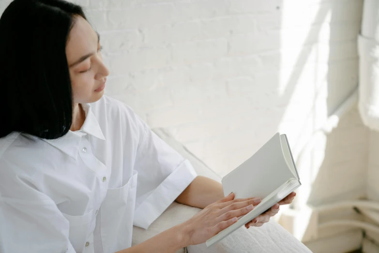 a woman sitting on a bed reading a book, a sketch, pexels contest winner, private press, wearing white shirt, korean woman, sleek hands, on a couch