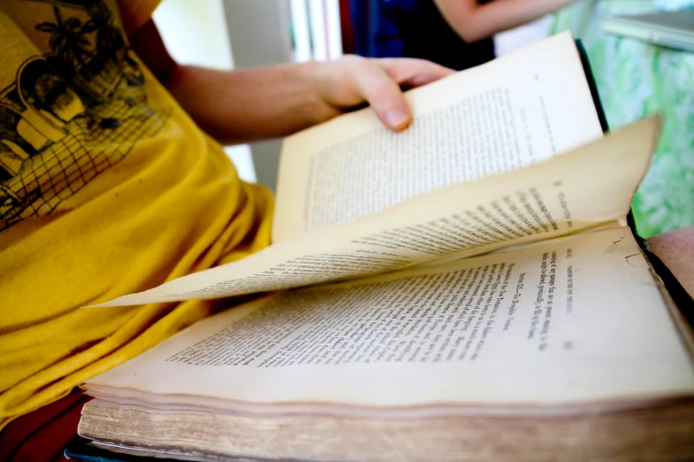 a person sitting on a bench reading a book, private press, over the shoulder closeup, holding a giant book, with one vintage book on a table, person in foreground