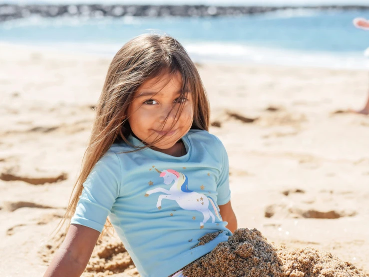 a little girl sitting on top of a sandy beach, a portrait, inspired by Charles Gleyre, unsplash, blue tight tshirt, unicorn, official product photo, with intricate detail