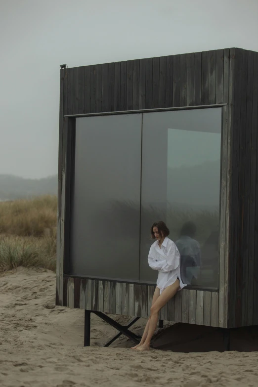 a woman sitting on a chair in front of a building, a picture, unsplash, conceptual art, stood outside a wooden cabin, under a gray foggy sky, beaching, nesting glass doors