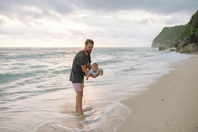 a man holding a baby on a beach, pexels contest winner, happening, avatar image, new zeeland, full body image, maintenance