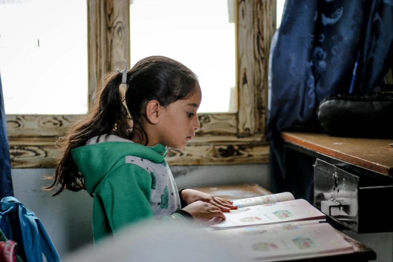 a little girl sitting at a desk reading a book, by Daniel Lieske, pexels contest winner, hurufiyya, from egypt, teaching, profile image, dressed in a worn