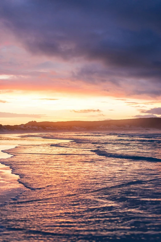 a man riding a surfboard on top of a sandy beach, during a sunset, wellington, which shows a beach at sunset, deep purple and orange