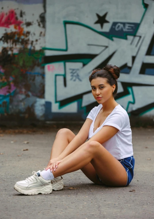 a woman sitting on the ground in front of a graffiti wall, inspired by Ion Andreescu, pexels contest winner, tan skin a tee shirt and shorts, wearing white sneakers, 5 0 0 px models, photo of a model