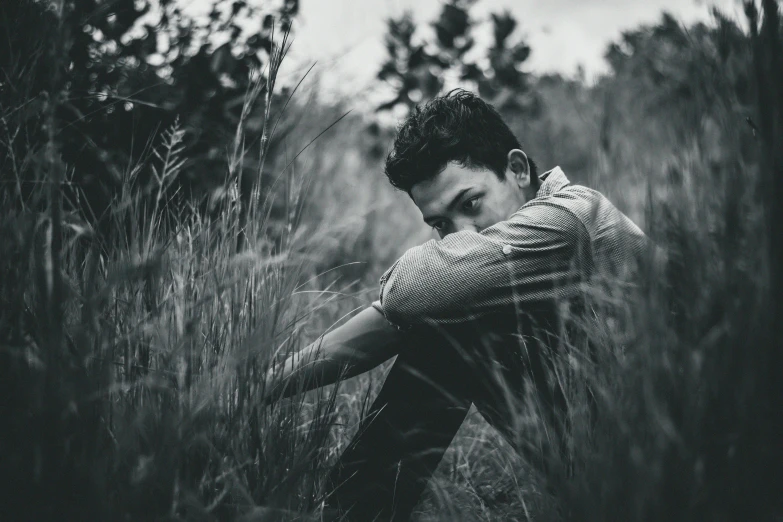 a man sitting in a field of tall grass, a black and white photo, by Adam Marczyński, pexels contest winner, realism, thoughtful pose, crouching, a handsome, over the shoulder