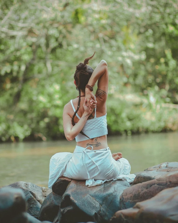 a woman sitting on top of a rock next to a river, armfold pose!, wearing white leotard, with a tree in the background, 2019 trending photo