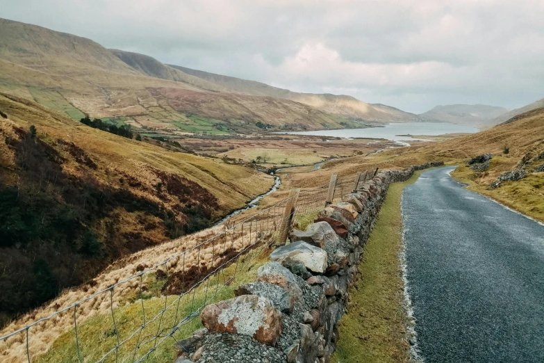 a road going down a hill next to a body of water, by Bedwyr Williams, pexels contest winner, stone wall, rolling foothills, thumbnail, leesha hannigan