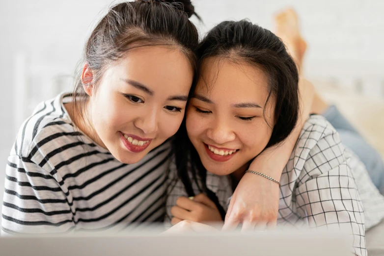 a couple of women laying on top of a bed, inspired by Wang Duo, trending on pexels, computer art, young cute wan asian face, looking at monitor, smiling playfully, south east asian with long