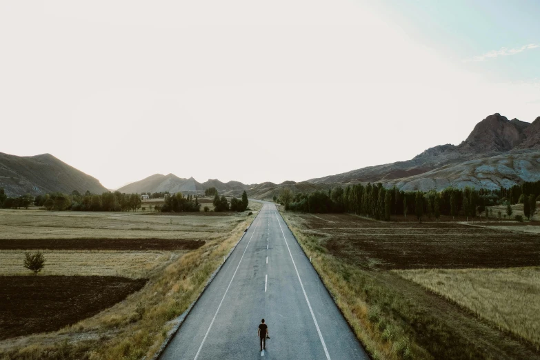 a person riding a bike down the middle of a road, a picture, unsplash contest winner, standing in a barren field, mountains in distance, drone photograpghy, walking down a street