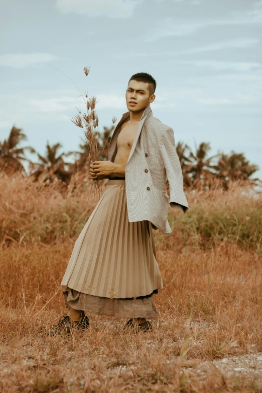 a man standing in a field holding a plant, an album cover, inspired by Byron Galvez, renaissance, pleated skirt, masculine appeal high fashion, indonesia, brown