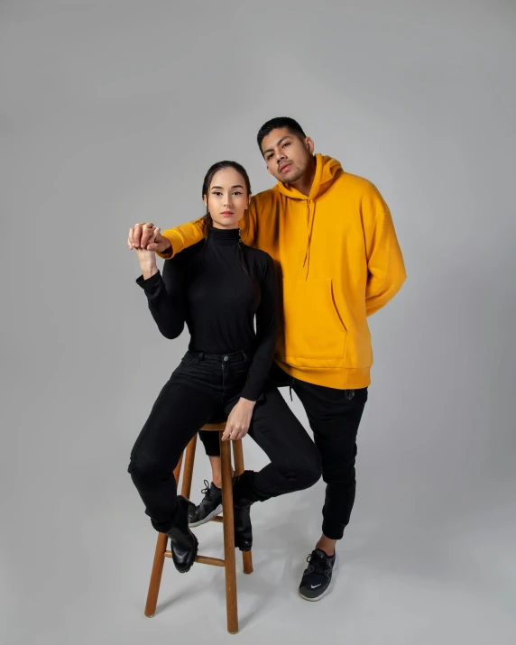 a man and a woman posing for a picture, wearing a yellow hoodie, wearing jeans and a black hoodie, profile image, sitting on a stool