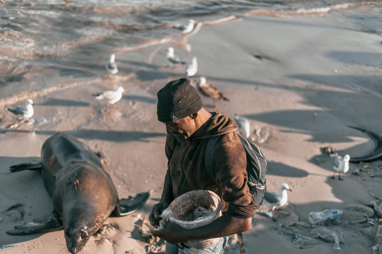 a man standing next to a sea lion on a beach, pexels contest winner, process art, offering a plate of food, working hard, indigenous man, south african coast