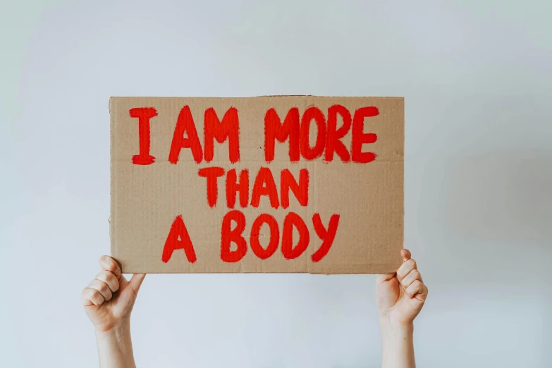 a person holding a sign that says i am more than a body, by Julia Pishtar, trending on pexels, anatomically incorrect, health supporter, cardboard, biological