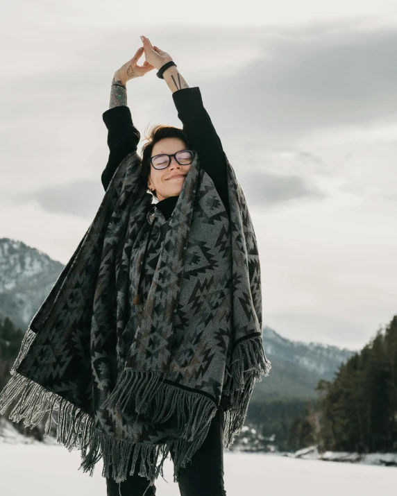 a woman standing on top of a snow covered field, an epic non - binary model, ornate poncho, hands in air, wearing black frame glasses