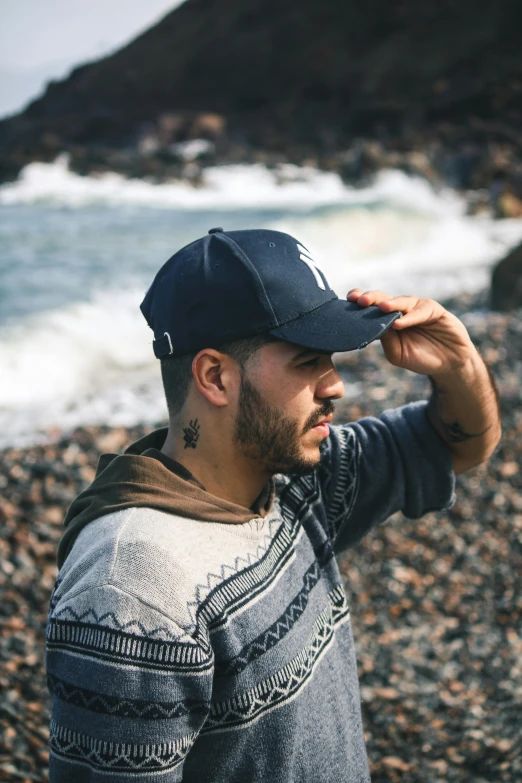 a man standing on a rocky beach next to the ocean, wearing a navy blue utility cap, beard stubble, streetwear, wearing a backwards baseball cap