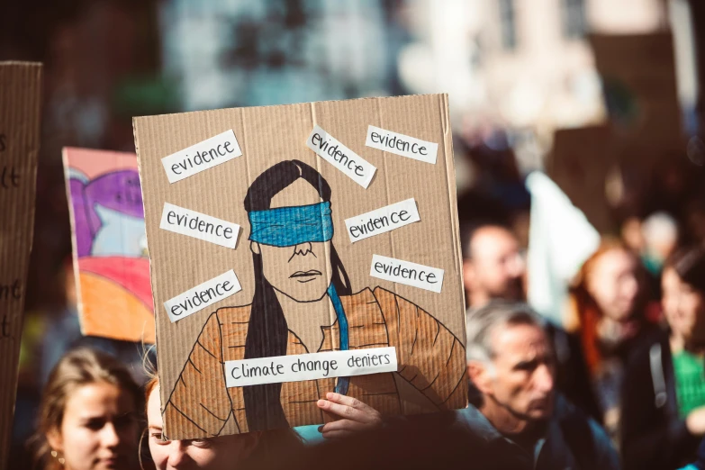 a person holding a cardboard sign in front of a crowd, a cartoon, by Emma Andijewska, trending on pexels, renaissance, blindfold, climate change, indigenous, science journal cover