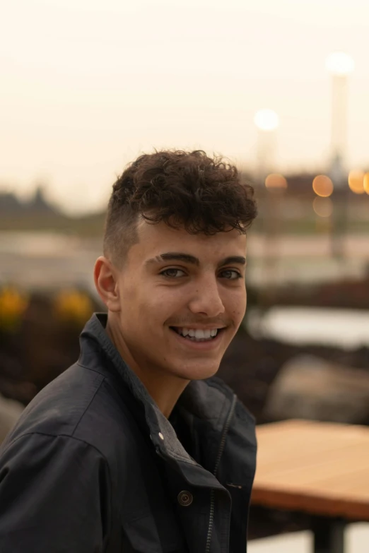 a young man is smiling at the camera, by Leo Michelson, unsplash, full color photograph, early evening, curly and short top hair, sam nassour