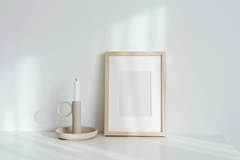 a picture frame sitting on top of a table next to a candle, a minimalist painting, trending on pexels, minimalism, light wood, white backdrop, morandi colour scheme, holding a candle holder