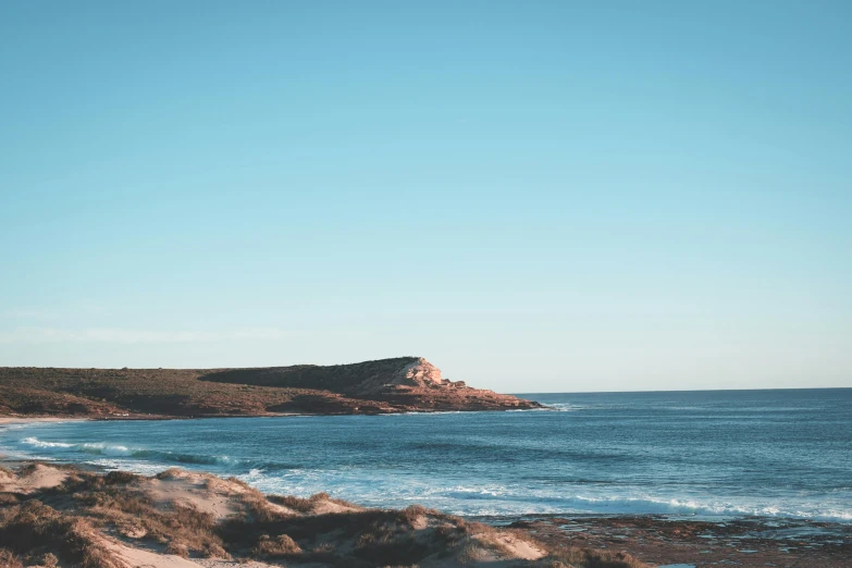 a man standing on top of a sandy beach next to the ocean, by Lee Loughridge, unsplash contest winner, minimalism, in the distance is a rocky hill, light blue clear sky, manly, seen from a distance