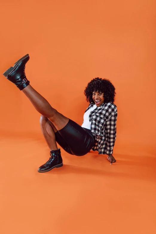 a woman sitting on the ground with her legs up, trending on pexels, black arts movement, in front of an orange background, leather boots, doing a sassy pose, giddy smirk