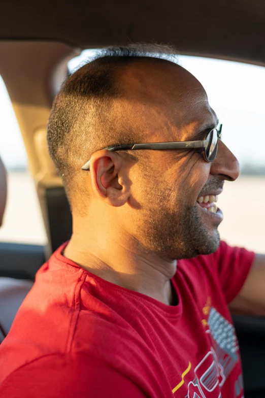 a man sitting in the driver's seat of a car, hurufiyya, smiling at each other, light stubble with red shirt, implanted sunglasses, in profile