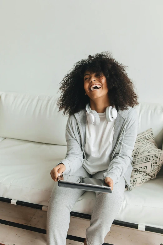 a woman sitting on a couch holding a tablet, pexels contest winner, happening, long afro hair, laughing your head off, wearing a track suit, white l shaped couch