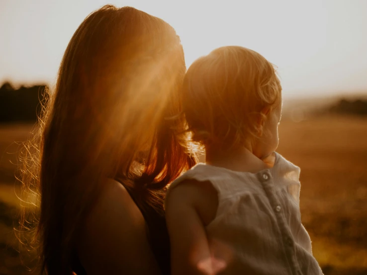 a woman holding a small child in a field, pexels contest winner, sun lit, looking outside, evening time, over the shoulder