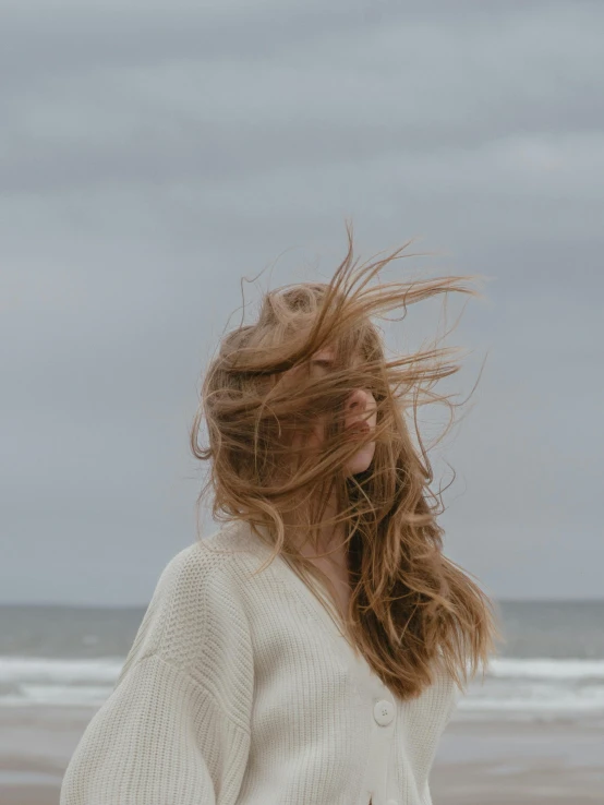 a woman standing on top of a sandy beach, inspired by Wilhelm Hammershøi, trending on pexels, long windy hair style, on a rainy day, a woman's face, wearing a white sweater