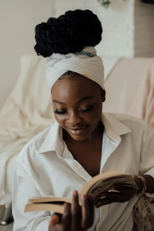 a woman sitting on a couch reading a book, by Chinwe Chukwuogo-Roy, pexels contest winner, afrofuturism, wearing white silk robe, hair styled in a bun, someone sits in bed, turning her head and smiling