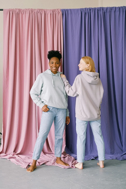 two women standing next to each other in front of a curtain, trending on pexels, wearing a hoodie and sweatpants, pastel color theme, wearing jeans, promotional image