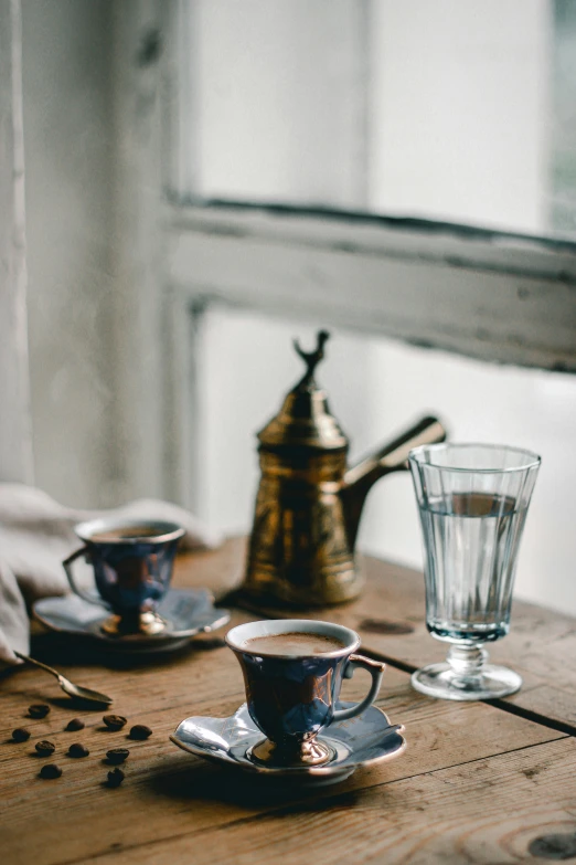 a wooden table topped with two cups of coffee, a still life, by Lucia Peka, trending on unsplash, renaissance, grey and blue theme, turkish and russian, soft window light, sparkling spirits