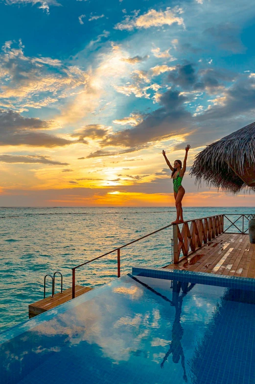 a woman standing on the edge of a swimming pool, which shows a beach at sunset, maldives in background, waving arms, dreampool rooms