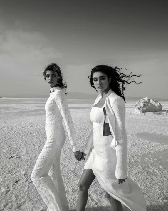 a couple of women walking across a sandy beach, a black and white photo, inspired by irakli nadar, trending on unsplash, surrealism, wearing futuristic white suit, :: madison beer, vera wang couture, on a desert road