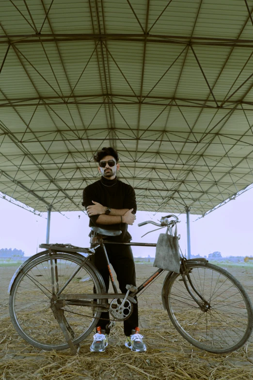 a man standing next to a bike in a field, an album cover, pexels contest winner, standing in an arena, india, ( ( theatrical ) ), shades