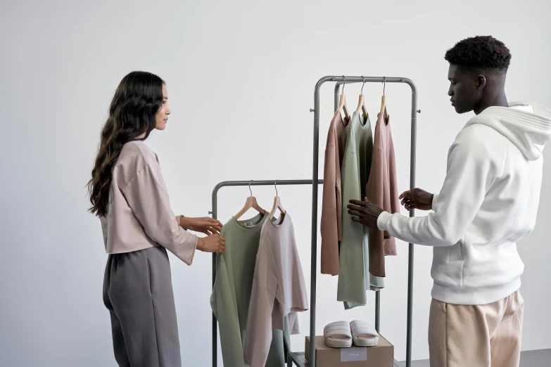 a man and a woman standing in front of a rack of clothes, muted colors with minimalism, presenting wares, collaborative, product display