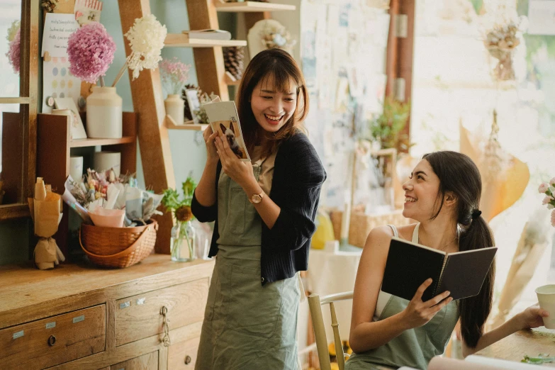 a woman standing next to a woman sitting at a table, pexels contest winner, happening, holding a book, flower shop scene, avatar image, smiling playfully