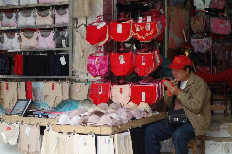 a man sitting in front of a display of underwear, wet market street, pink and red color scheme, red cap, red bra