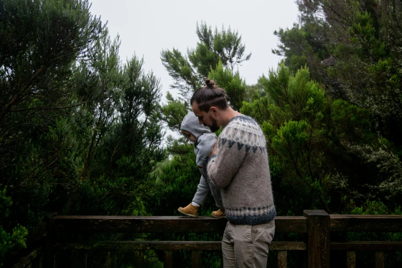 a man holding a small child in his arms, by Jessie Algie, unsplash, kahikatea, standing on a bridge, maritime pine, sweater