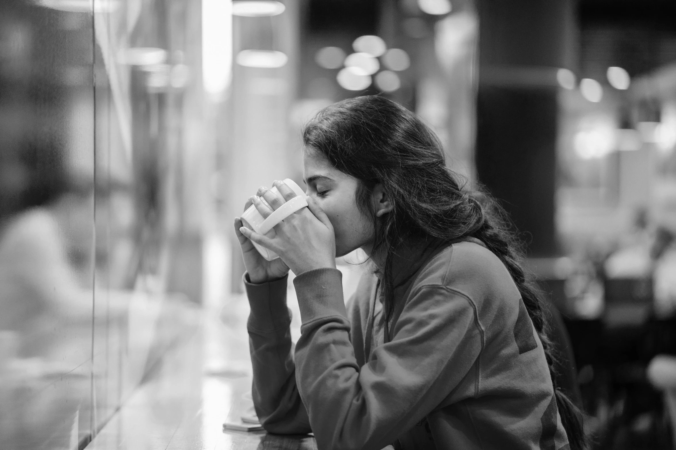 a woman sitting at a table with a cup of coffee, a black and white photo, pexels, romanticism, in a mall, hand over mouth, teenager girl, young middle eastern woman