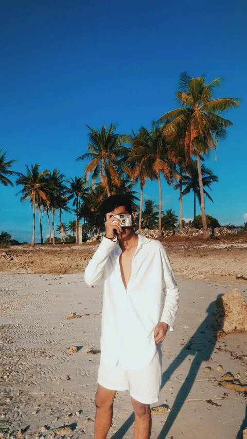 a man standing on a beach talking on a cell phone, a polaroid photo, unsplash, sumatraism, wearing white pajamas, cai xukun, palm trees, posing for a picture