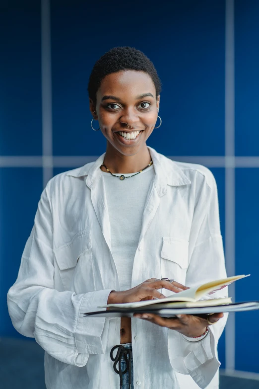 a woman standing in front of a blue wall holding a clipboard, academic art, dark skinned, textbooks and books, wearing white robe, wearing business casual dress