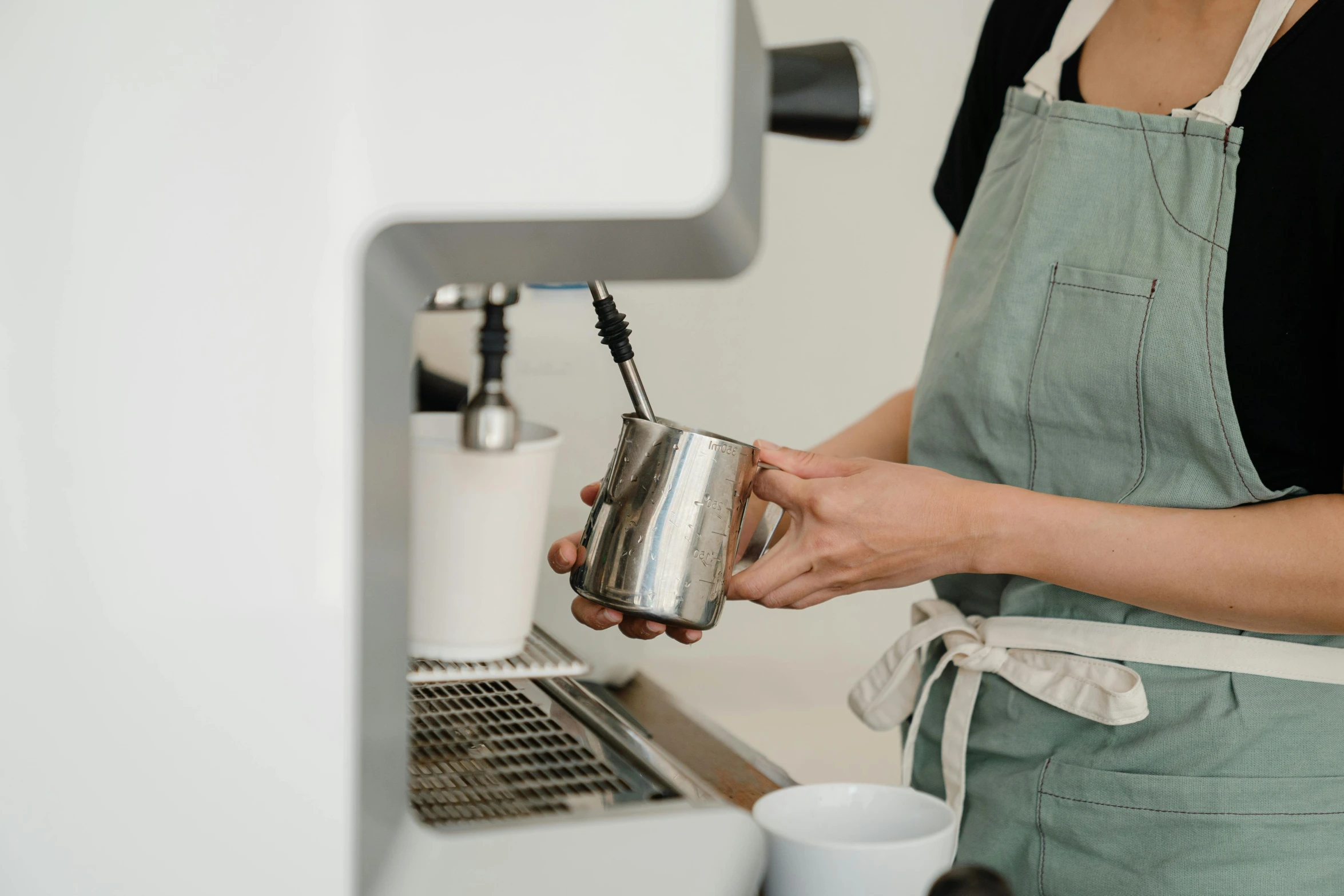 a woman standing in front of a coffee machine, pexels contest winner, white apron, immaculate detail, lachlan bailey, stainless steel