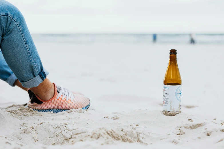 a person sitting on a beach next to a bottle of beer, unsplash contest winner, running shoes, dezeen, australian beach, paddy boehm