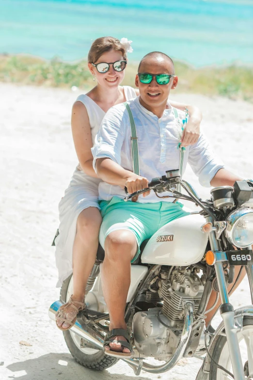 a man and woman riding on the back of a motorcycle, sea - green and white clothes, white sand, wedding photo, both smiling for the camera