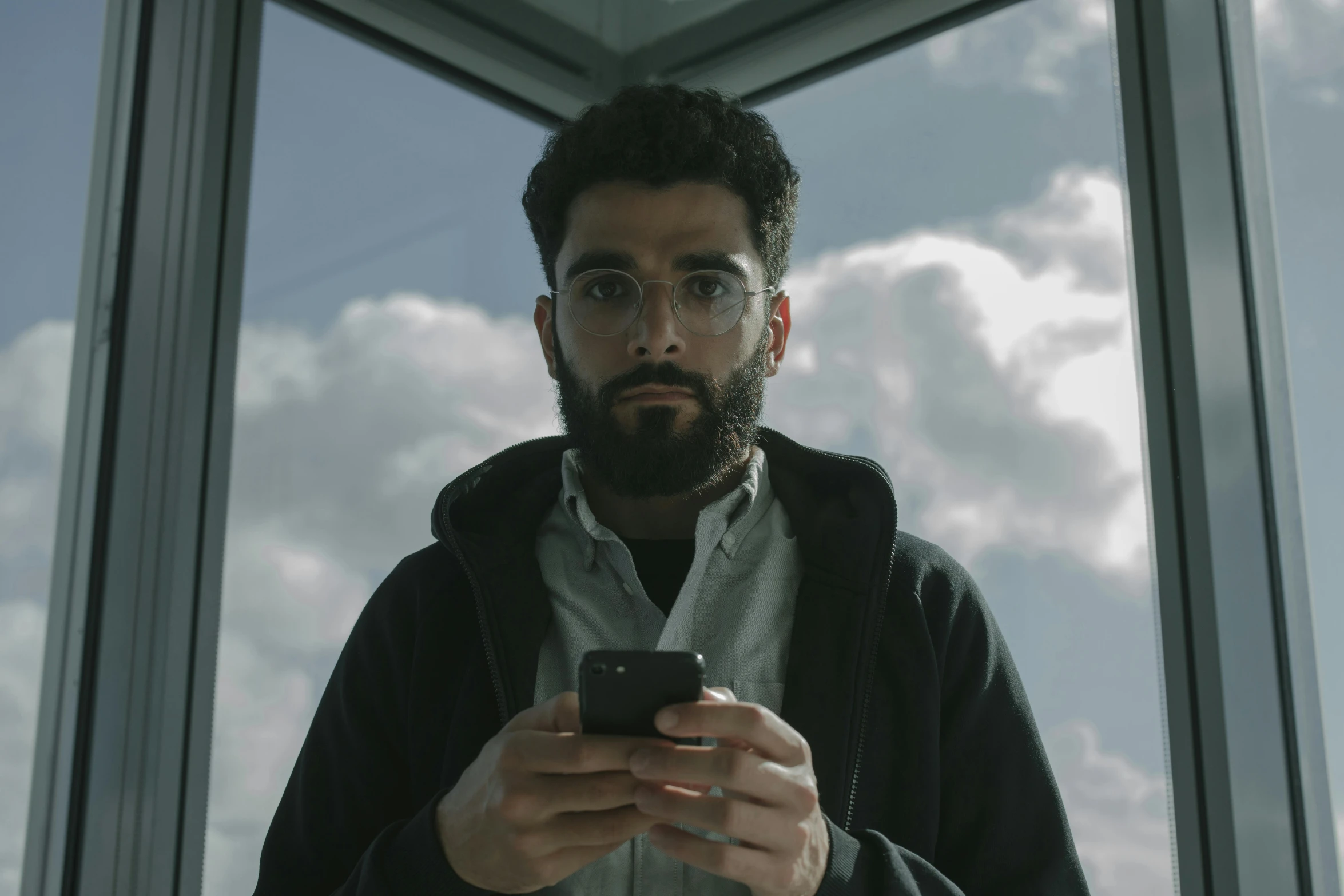 a man standing in front of a window looking at his cell phone, pexels contest winner, serial art, a portrait of rahul kohli, from a 2 0 1 9 sci fi 8 k movie, jewish young man with glasses, cloudy day