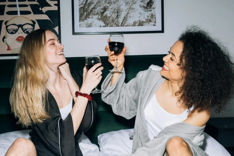 a couple of women sitting on top of a bed holding wine glasses, cosy vibes, dark grey robes, very excited, abcdefghijklmnopqrstuvwxyz