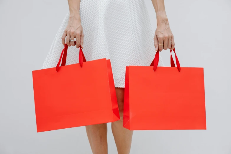 a woman in a white dress holding two red shopping bags, by Nicolette Macnamara, pexels contest winner, minimalism, coral red, short skirt length, luxury brand, round-cropped