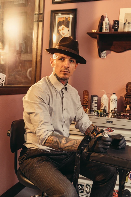 a man sitting at a desk with a tattoo machine, a tattoo, inspired by Jeremy Henderson, black stetson hat, looking confident, owen gent, hairless