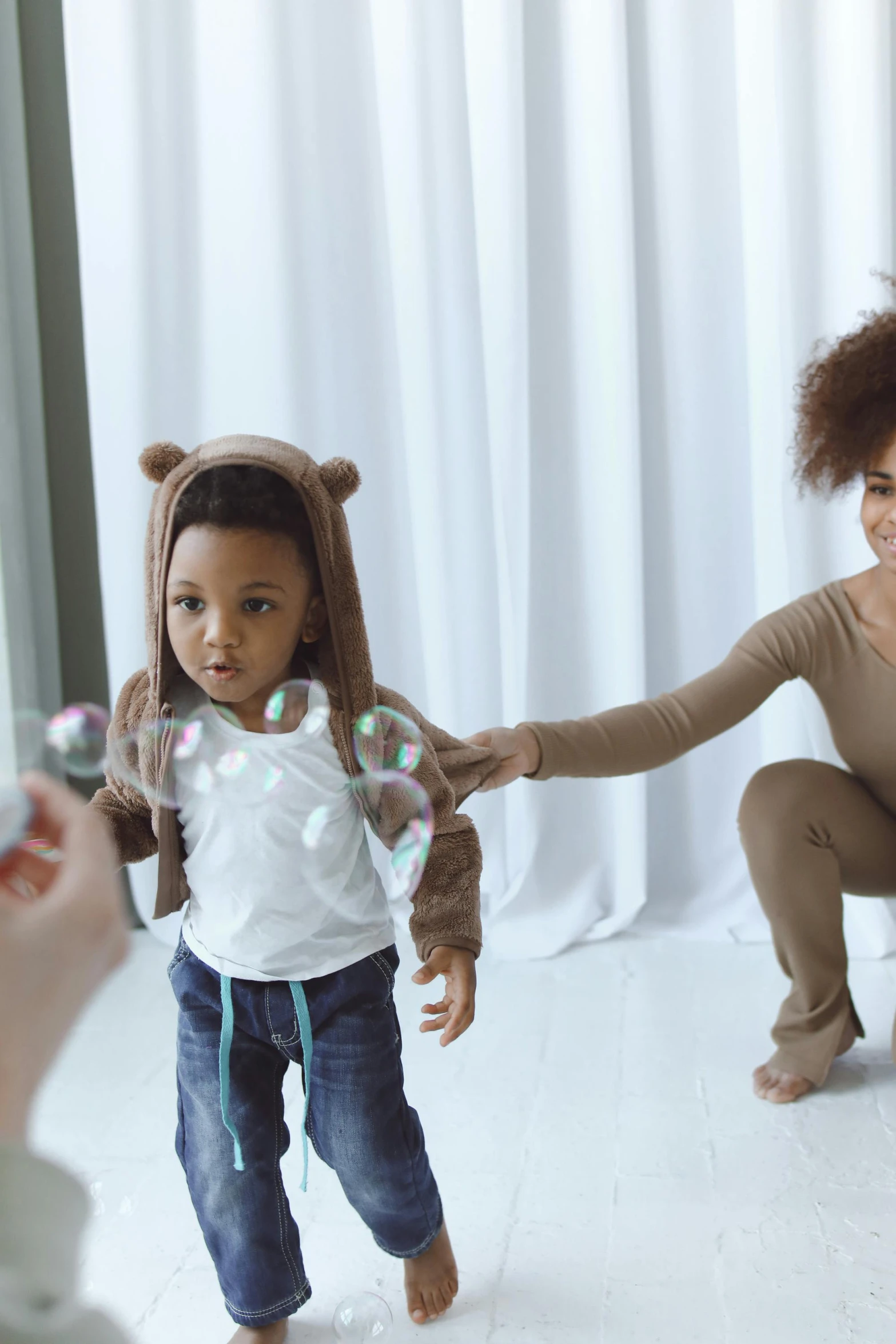 a woman and a child are playing with soap bubbles, by Arabella Rankin, interactive art, holding a teddy bear, with afro, running towards camera, indoor scene