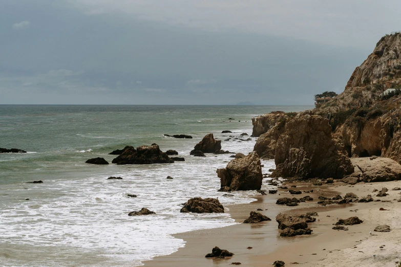 a man standing on top of a sandy beach next to the ocean, rocky cliff, overcast day, malibu canyon, background image
